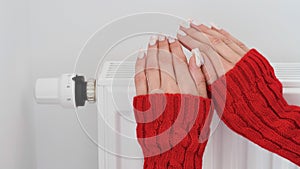 Close-up of a woman in red sweater warms her hands over the battery on background of white wall.Macro photo of woman`s hands,copy
