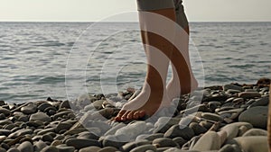 Close-up of a woman's bare feet with their pants rolled up walking on a rocky beach. The sea is splashing in the
