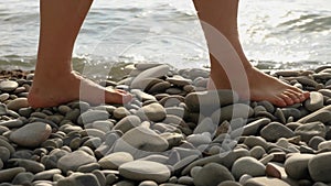 Close-up of a woman's bare feet walking along a stone beach. The sea is splashing in the background. The camera follows