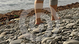 Close-up of a woman's bare feet walking on the rocks by the sea. Dry seaweed and water in the background.
