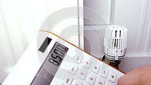 Close-up of a woman`s hand holding a calculator near radiator, temperature regulation in house with thermostat on white radiator,