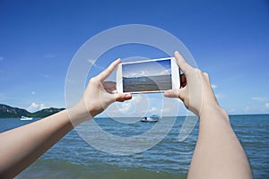 Close up of woman's hand holding smart phone, mobile, smart phone over blurred beautiful blue sea and fishing boat