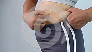 Close-up of woman`s hand pinching excessive belly fat isolated on gray background. Woman fat belly. Obesity and Overweight