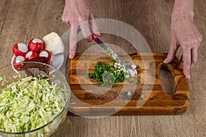 Close-up of woman's hands chopping green onions with a kitchen knife for making salad on a wooden cutting board.