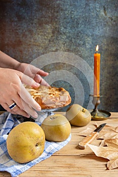 Close-up of woman`s hands holding an apple pie on wooden table with pippin apples and candle,