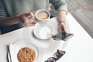 Close-up of woman`s hands holding cell phone while drinking coffee and eating oat cookie in a modern cafe.