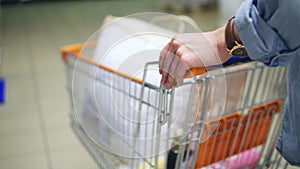 Close up of a woman`s hands holding and pushing a trolley with goods in the supermarket. Backside view