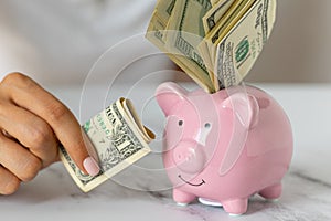 Close-up of a woman\'s hands put dollar paper money in a piggy bank, next to a calculator on the table. economy, crisis and