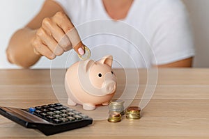 Close-up of a woman's hands put a euro coin in a piggy bank, next to a calculator on the table.saving money in a crisis and