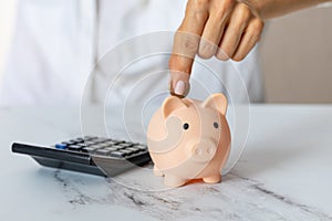 Close-up of a woman's hands put a euro coin in a piggy bank, next to a calculator on the table.saving money in a crisis and