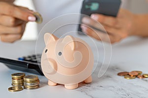 Close-up of a woman\'s hands put a euro coin in a piggy bank, next to a calculator on the table.saving money in a crisis and