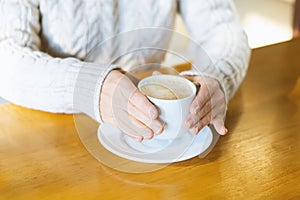 Close-up of a woman`s hands in a warm sweater holding a white Cup of hot coffee, chocolate or tea in a coffee shop against the bac