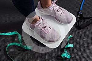 Close up woman`s sneakers stand on an electric scale on a black floor. Measuring tape and exercise rope near