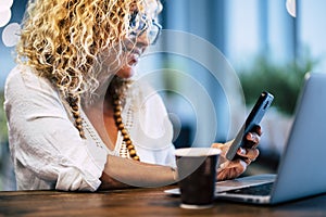 Close up of woman smiling and looking at her phone with her laptop and coffee - beautiful and worker adult lady using and working