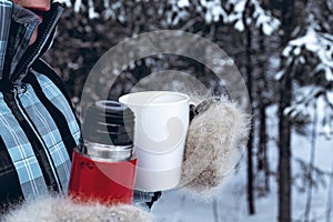 Close-up. Woman in a warm clothing holds a red thermos with a hot drink and a white mug. Winter forest background