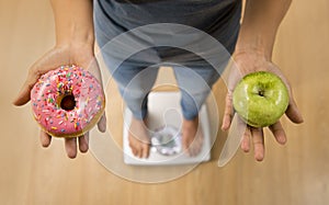 Close up woman on weight scale holding in her hand apple fruit and donut as choice of healthy versus unhealthy food