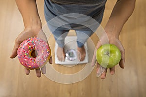 Close up woman on weight scale holding in her hand apple fruit and donut as choice of healthy versus unhealthy food