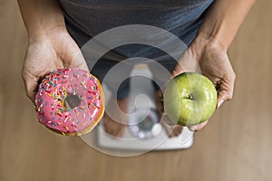 Close up woman on weight scale holding in her hand apple fruit and donut as choice of healthy versus unhealthy food