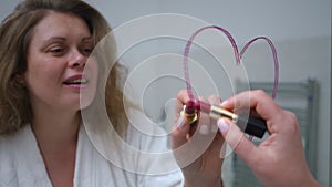 Close-up. A woman in a white bathrobe smiles and draws a heart with red lipstick on the mirror while standing in the