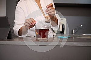 Close-up woman in white bathrobe, sweetening his tea, standing at kitchen table with open digital tablet, in a