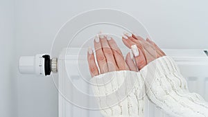 Close-up of a woman in white sweater warms her hands over the battery on background of white wall.Macro photo of woman`s hands,