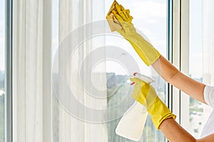 Close up of woman in yellow rubber gloves cleaning window with cleanser spray and yellow rag at home or office, copy space.