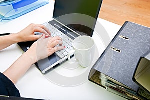 Close up of womans hands typing on laptop with folders and mug