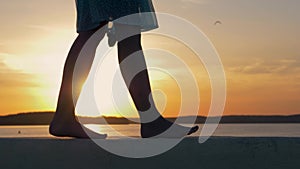 Close-Up Of Women Feet Are On The Beach In The Flicker Rays Of The Sunset