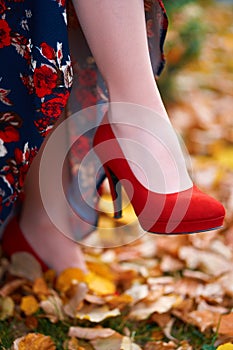 Close up of women`s feet shod in red high heels shoes, autumn season, yellow fallen leaves as background