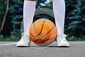Close up wonan`s feet in white sneakers and whote long socks, with a ball on a basketball court.