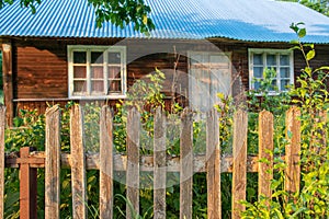 Close up on wooden fence standing in front of old, wooden village house. Building surrounded by trees and bushes on a summer day