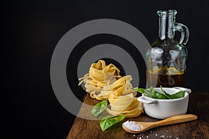Close-up of wooden spoon with salt, basil in bowl, bottle with olive oil and raw tagliatelle, on wooden table, black background