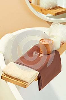 Close-up of a wooden tray placed across the bathtub with cloth towels, pink salt, scented candles and a bath sponge. Space with