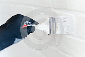 Close-up of a worker& x27;s hand with a brush painting a brick wall with white paint.