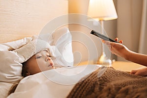 Close-up of worried mother sitting on bed beside sick daughter with high fever and calling on phone to family doctor.