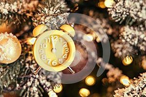 Close-up of a yellow clock showing the countdown to twelve o`clock hangs on the Christmas tree next to the glowing golden lights