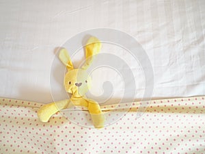 Close up yellow rabbit doll laying on white bed under cream blanket pink polka dot pattern