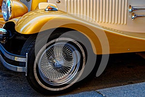 A close-up of a yellow vintage cars front side, showcasing the detailed wire-spoke wheel, chrome bumper, and sleek body design