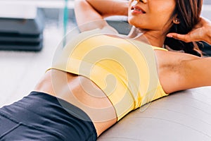 Close-up of young active and fitness Asian woman doing sit ups and crunches inside gym with exercise ball in background