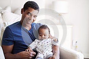 Close up of young adult African American  father sitting in an armchair holding his three month old baby son, smiling to camera, f