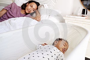 Close up of young adult mother lying on her bed looking down at her three month old baby sleeping in his cot