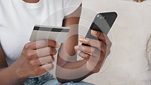 Close-up young African American female shopper holding smartphone and credit card in hands using instant easy mobile