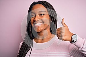 Close up of young african american woman wearing pink sweater over isolated background doing happy thumbs up gesture with hand