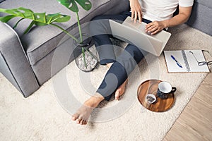 Close up of young Asian man sitting on floor working from home using laptop computer and notebook. Work from home concept