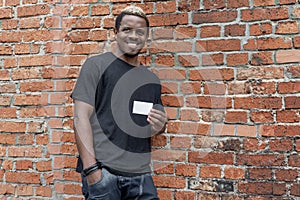 Dark-skinned man in T-shirt showing blank business card on bricked background.