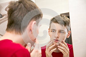 Close up of young attractive man with problematic skin and scars from acne looks in the mirror in the bathroom