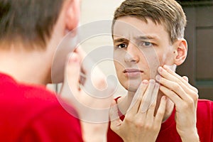 Close up of young attractive man with problematic skin and scars from acne looks in the mirror in the bathroom