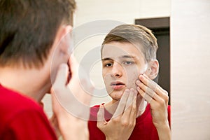 Close up of young attractive man with problematic skin and scars from acne looks in the mirror in the bathroom