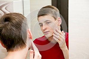 Close up of young attractive man with problematic skin and scars from acne looks in the mirror in the bathroom