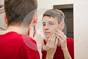 Close up of young attractive man with problematic skin and scars from acne looks in the mirror in the bathroom